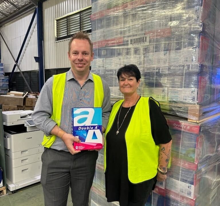A man holding a ream of paper stands next to a woman; both are wearing high-visibility safety vests