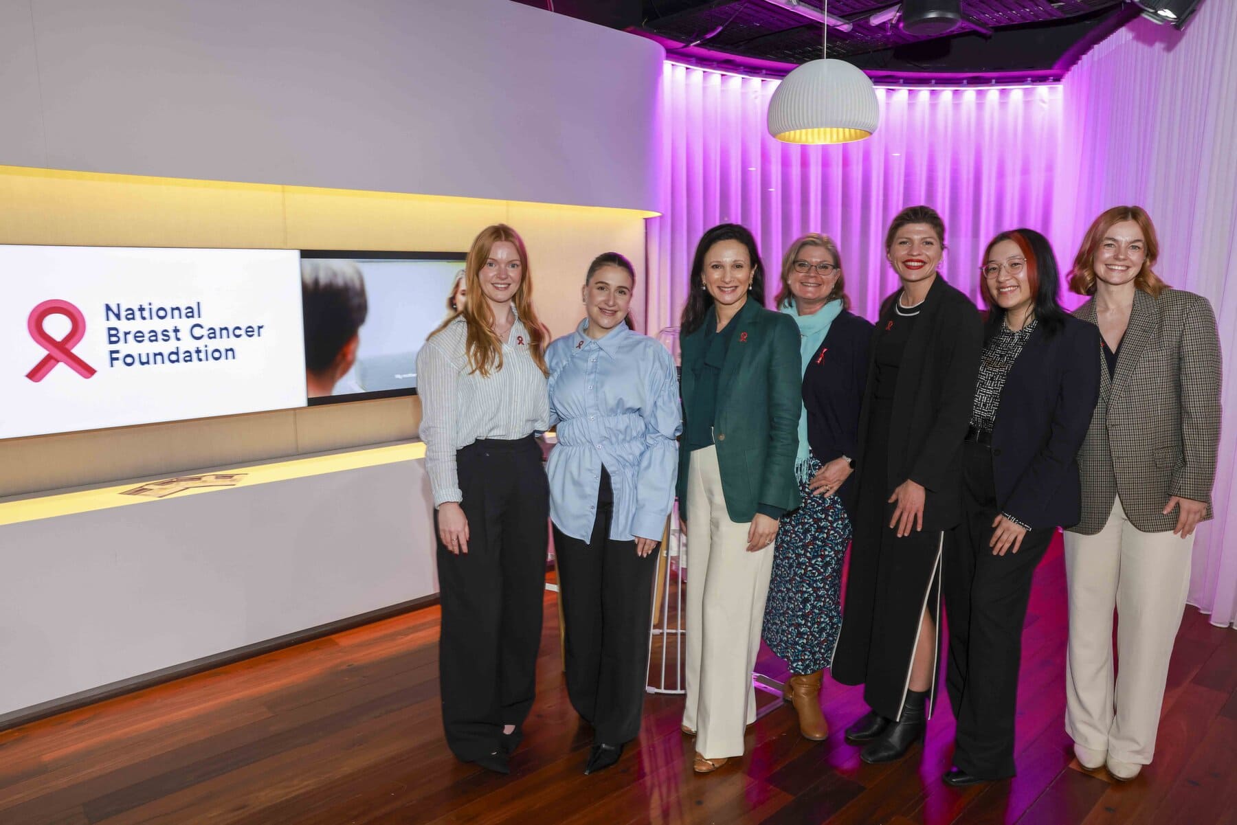 Seven people from the National Breast Cancer Foundation (NBCF) Team smiling next to the NBCF logo
